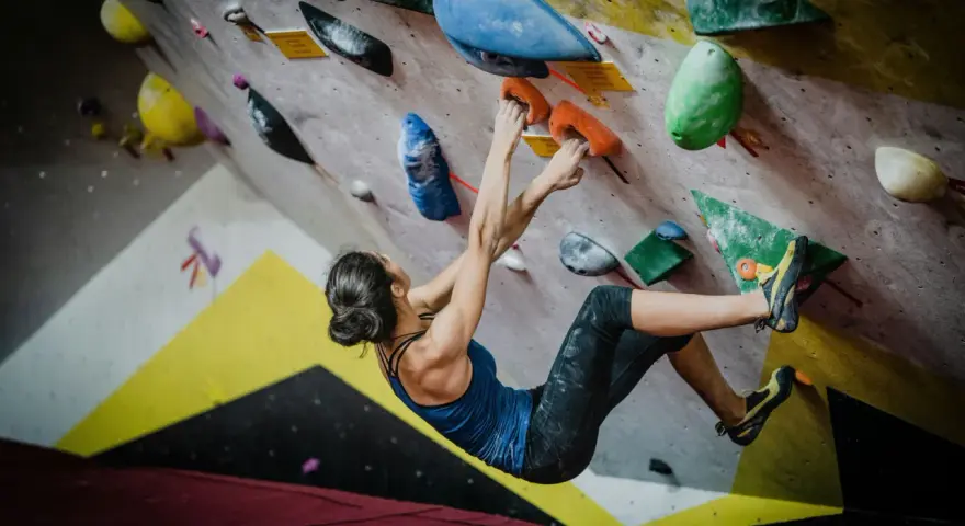 Kobieta wspina się po kolorowej ścianie, szukając chwytów. To właśnie bouldering – sportowa wspinaczka bez liny, na niskich ścianach.