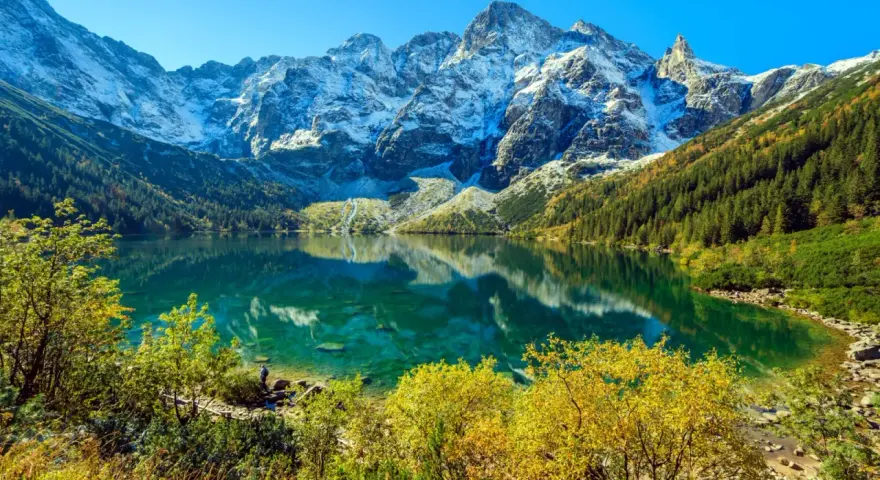 Turkusowe Morskie Oko odbija ośnieżone szczyty Tatr. Jesienne drzewa otaczają taflę wody.