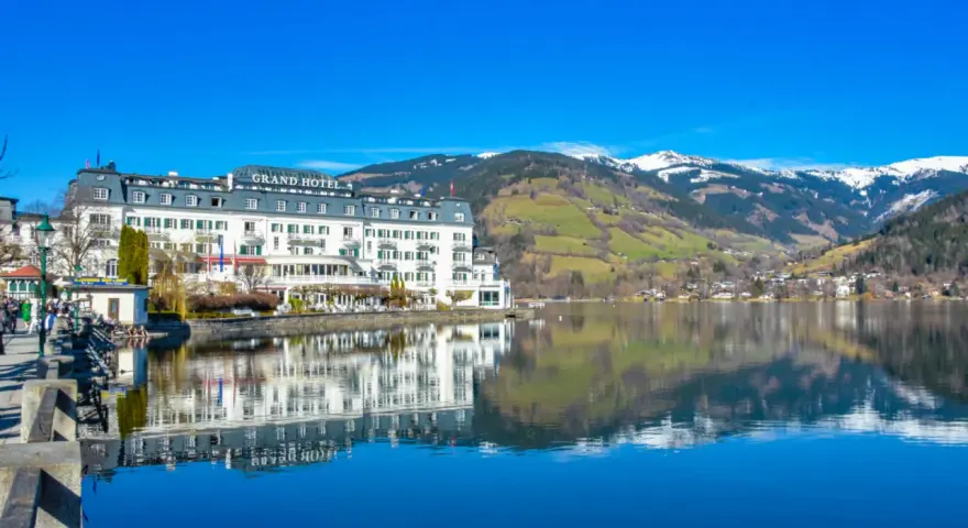 Grand Hotel nad jeziorem w Zell am See, z odbiciem w spokojnej wodzie i ośnieżonymi górami w tle.