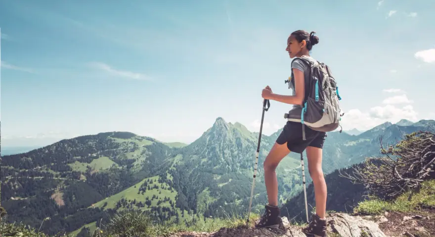 Młoda kobieta z plecakiem i kijkami trekkingowymi podziwia górski krajobraz. Pokazuje, jak chodzić z kijkami trekkingowymi, by czerpać radość z wędrówki.