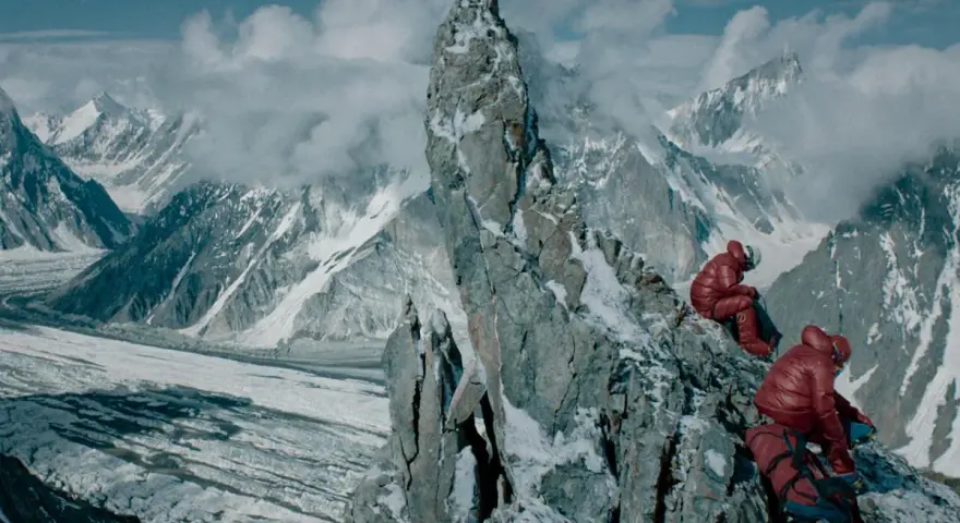 Dwóch alpinistów w czerwonych kurtkach odpoczywa na skalistym szczycie, podziwiając lodowcową dolinę i ośnieżone góry.