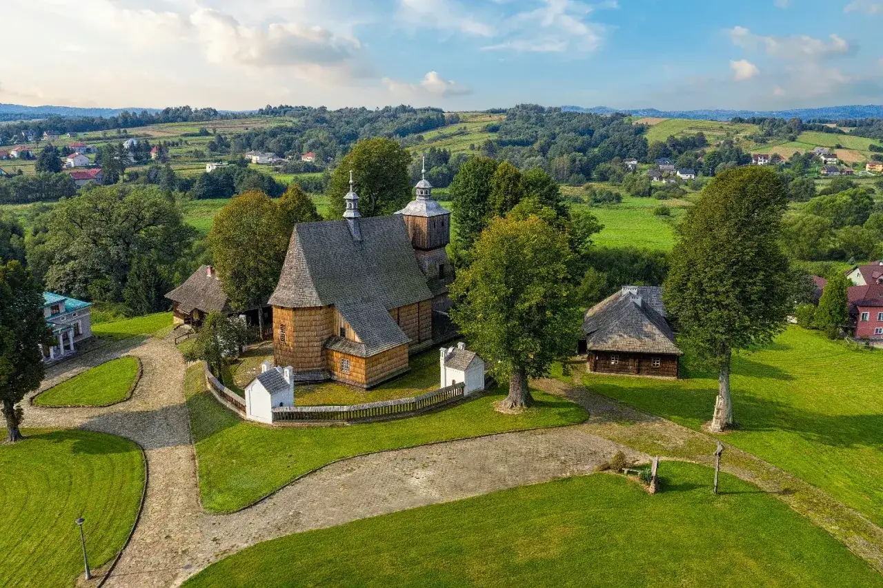 Drewniana cerkiew i zabudowania tworzą malowniczy krajobraz w Beskidzie Niskim. Wioska otoczona zielenią i pag&oacute;rkami.