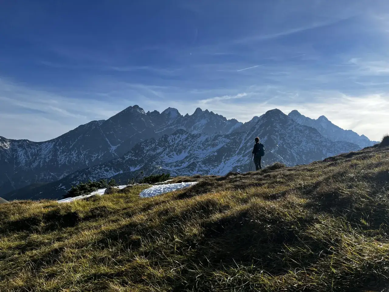 Osoba z plecakiem i kijkami maszeruje po trawiastym zboczu. W tle majestatyczne Tatry Bielskie, pokryte śniegiem.