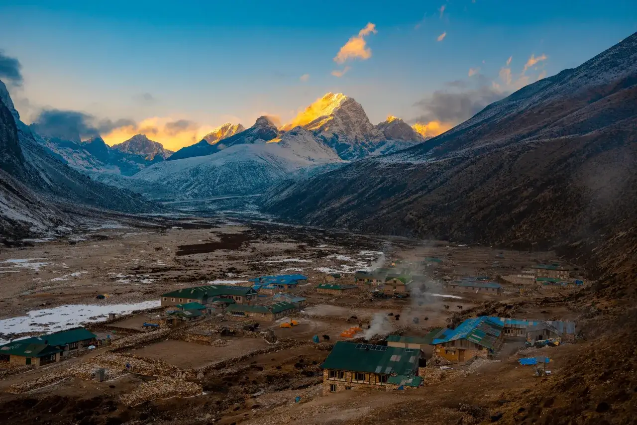 Wioska u podnóża ośnieżonych szczytów. Dym unosi się z domów, sugerując ciepło i życie. Planujesz wyprawę? Sprawdź Everest Base Camp koszty.