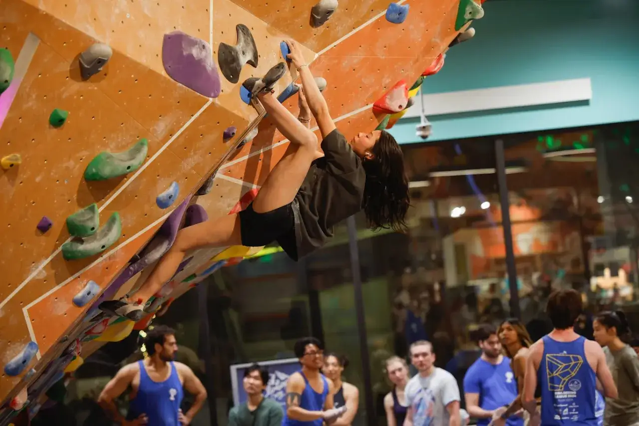 Kobieta wspina się po kolorowej ścianie w hali do boulderingu. To świetny sposób na aktywność fizyczną, pokazujący, co to bouldering.