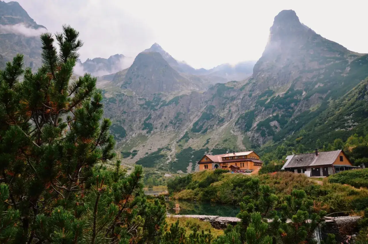 Widok na Tatry od strony słowackiej. Dwa schroniska górskie nad jeziorem, otoczone zielenią i skalistymi szczytami w chmurach.