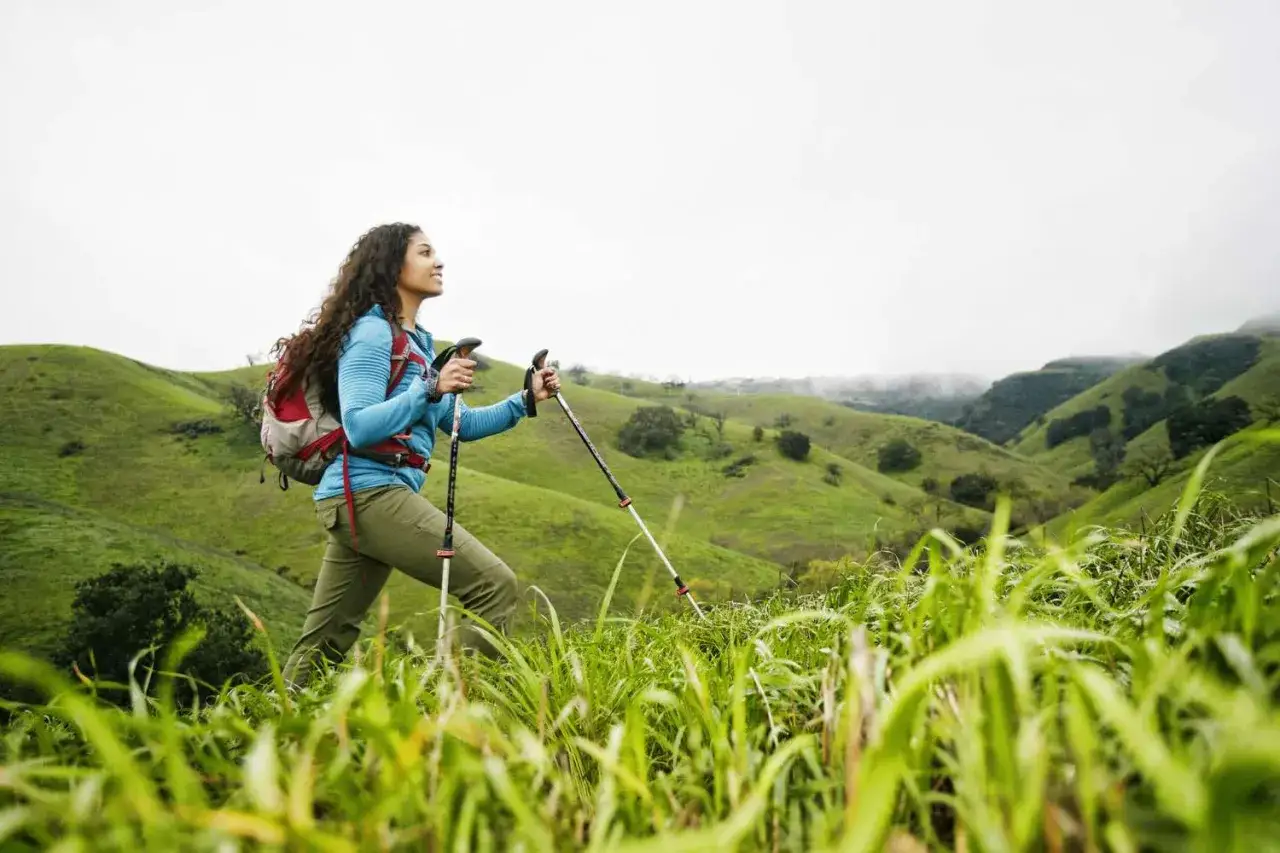 Kobieta z plecakiem i kijkami trekkingowymi podziwia zielone wzgórza. Pokazuje, jak chodzić z kijkami trekkingowymi, czerpiąc radość z wędrówki.