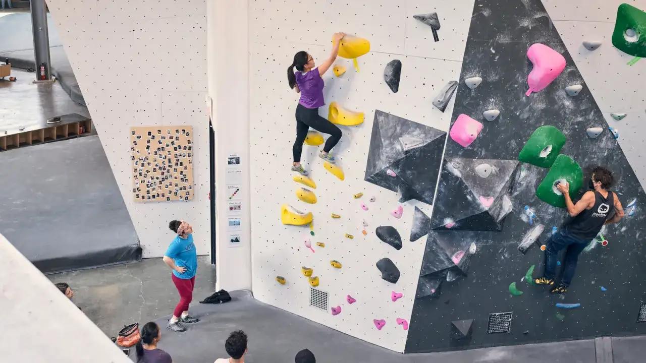 Bouldering co to? Wspinaczka po niskich ściankach bez lin, z kolorowymi chwytami. Na zdjęciu widać osoby wspinające się i obserwujące.