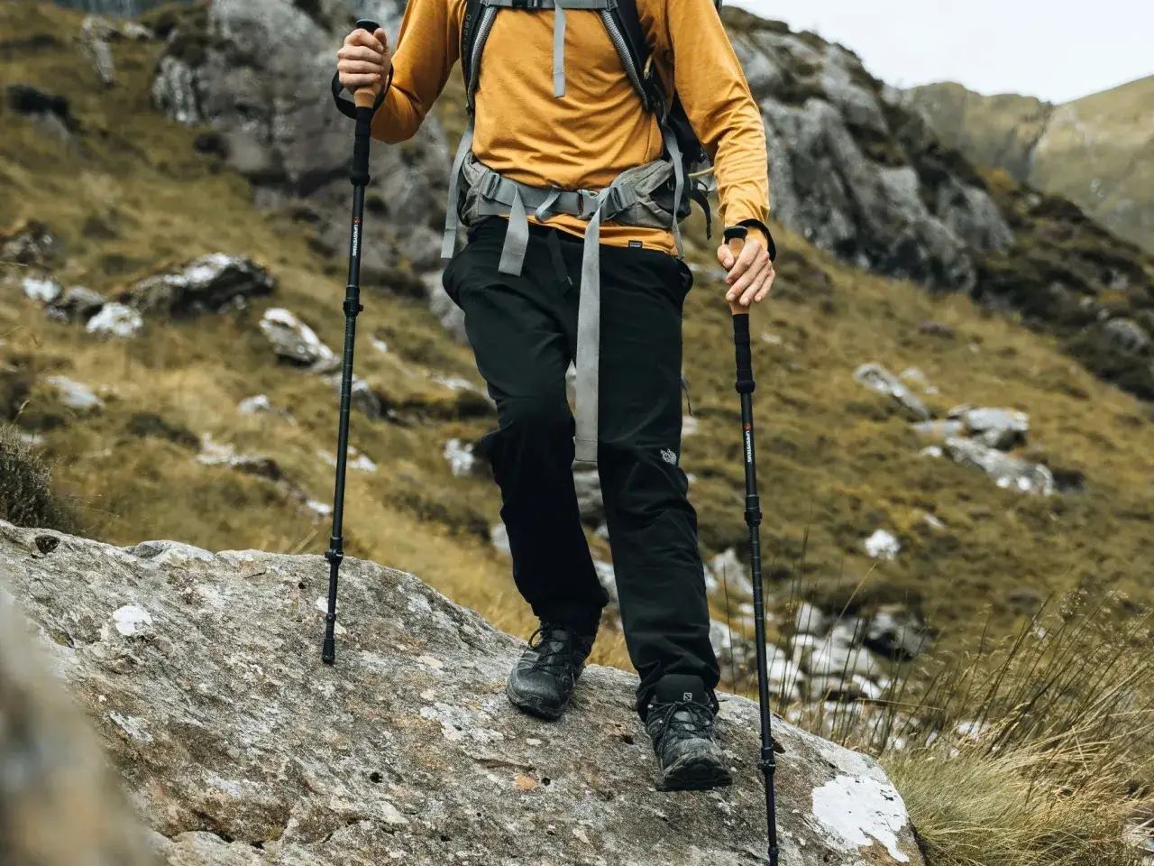 Osoba w żółtej bluzie i czarnych spodniach z kijkami trekkingowymi pokonuje kamienisty szlak. To świetny przykład, jak chodzić z kijkami trekkingowymi.