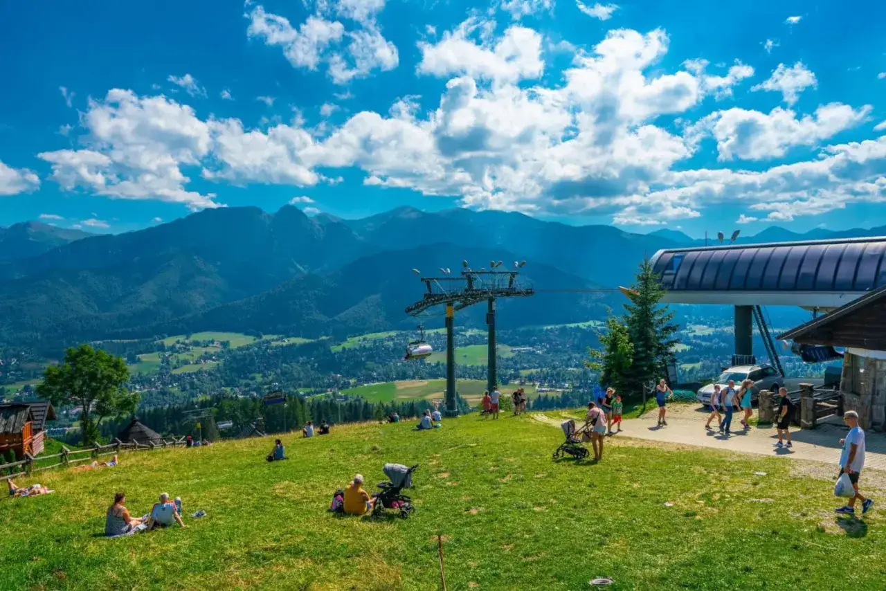 Widok na Tatry z Gubałówki. W oddali widać góry, a na pierwszym planie zieloną trawę, ludzi odpoczywających i wyciąg krzesełkowy na Gubałówkę.