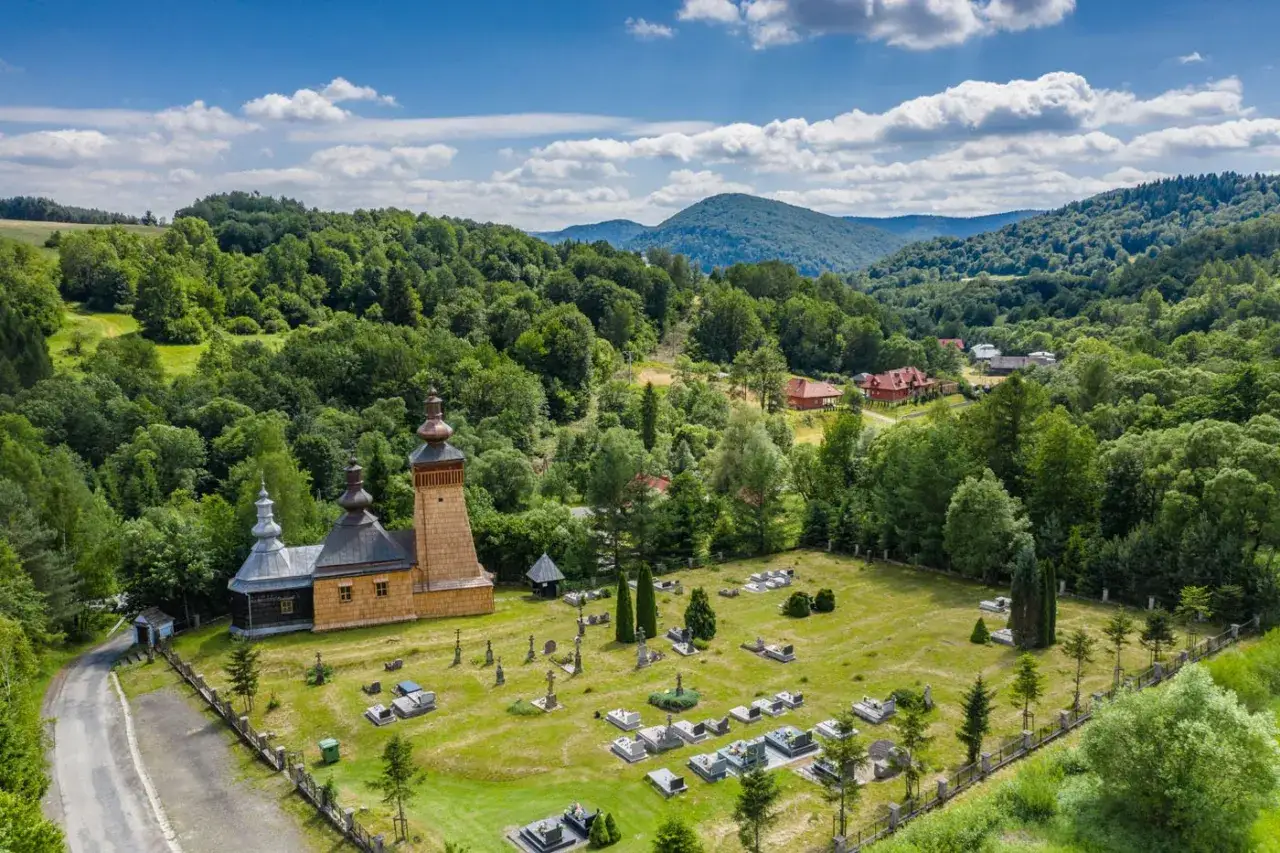 Drewniana cerkiew i cmentarz w malowniczej miejscowości Beskid Niski. W tle zielone wzg&oacute;rza i domy.