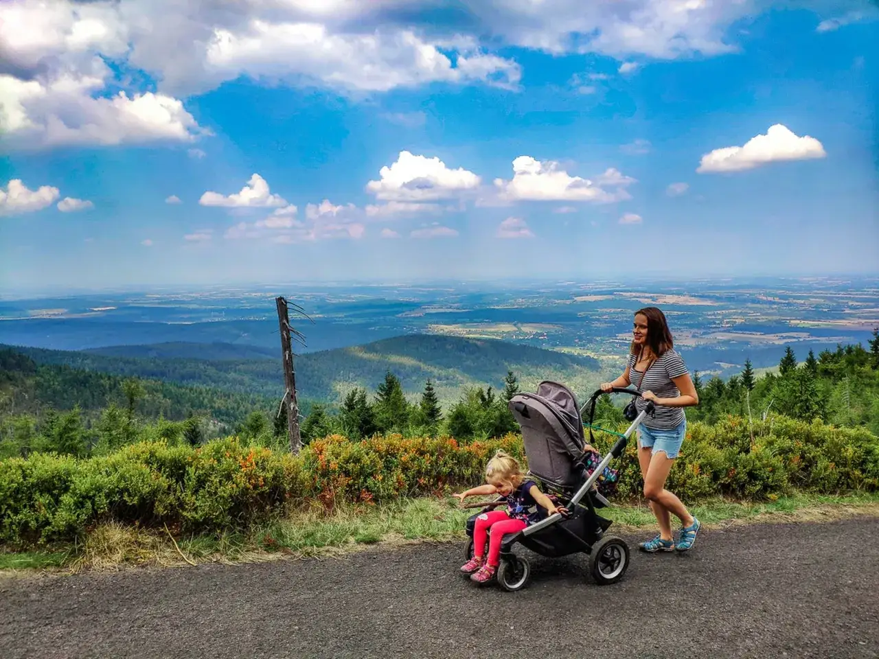 Kobieta pcha wózek z dzieckiem po górskiej ścieżce, podziwiając rozległy krajobraz. Tatry z wózkiem to piękna przygoda.