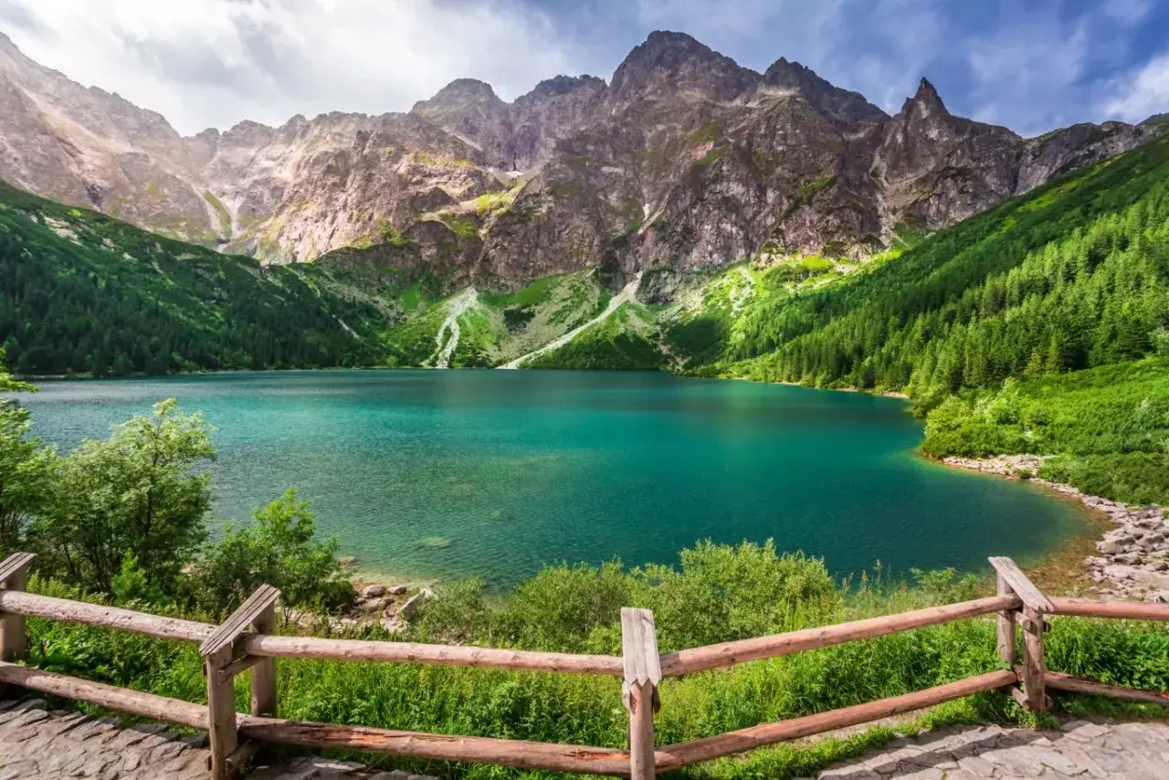 Turkusowe jezioro otoczone zielonymi zboczami g&oacute;r, przypominające beskidzkie Morskie Oko. Drewniane ogrodzenie prowadzi do malowniczego widoku.