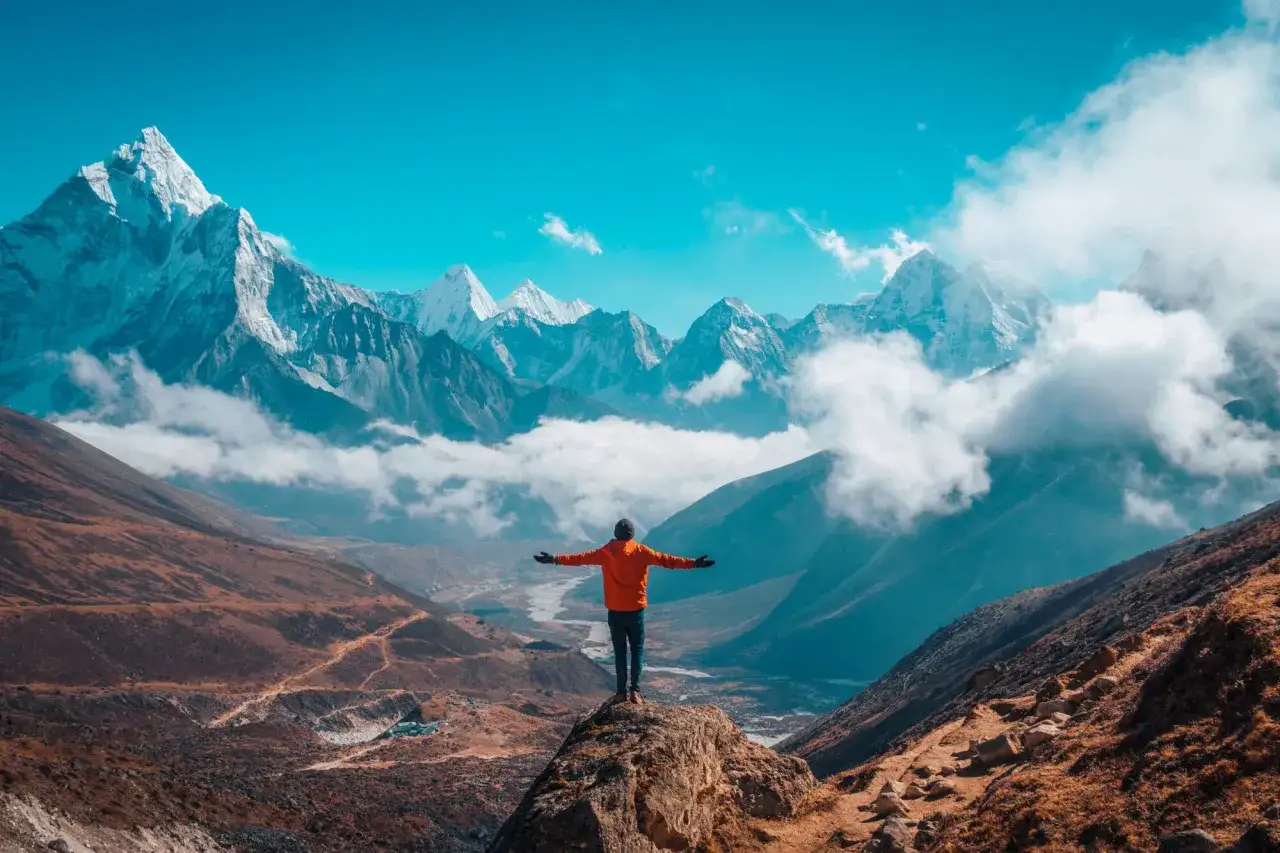 Widok na majestatyczne góry Himalajów, człowiek na szczycie skały z rozłożonymi rękami. Podróż na Everest Base Camp, poznaj jej koszty.