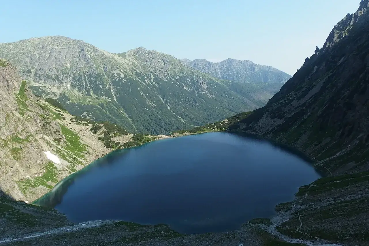 Głęboki, granatowy staw pod Rysami, otoczony stromymi, skalistymi zboczami g&oacute;r porośniętych zielenią.