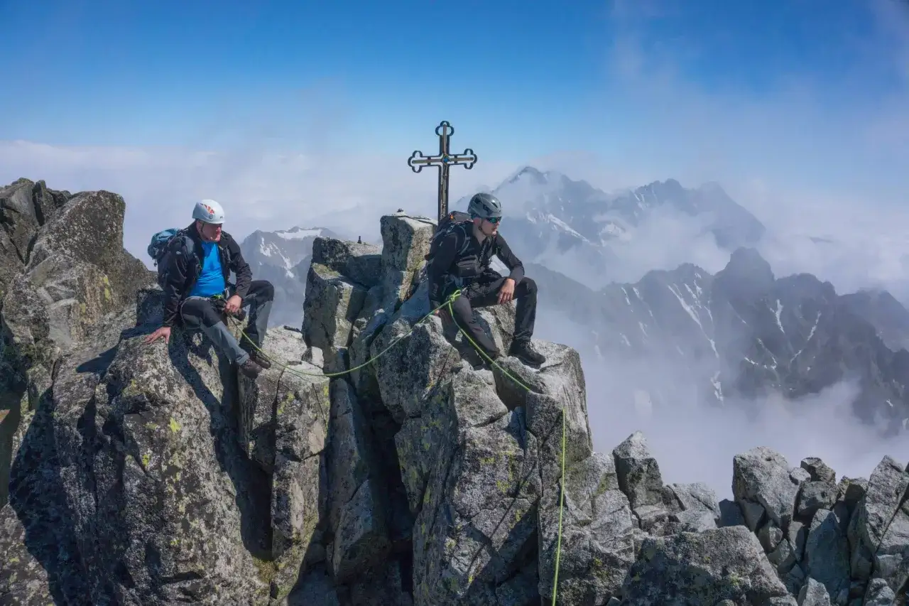 Dwóch alpinistów odpoczywa na szczycie. W tle rozciąga się majestatyczny widok z polski na Tatry Wysokie, z charakterystycznym krzyżem.