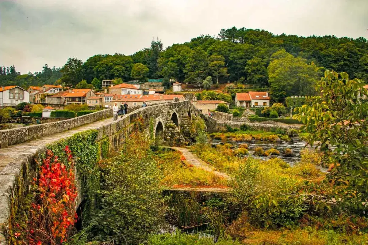 Kamienny most, porośnięty bluszczem i jesiennymi liśćmi, stanowi część malowniczych tras Camino de Santiago. W tle widać urokliwą wioskę i bujną zieleń.
