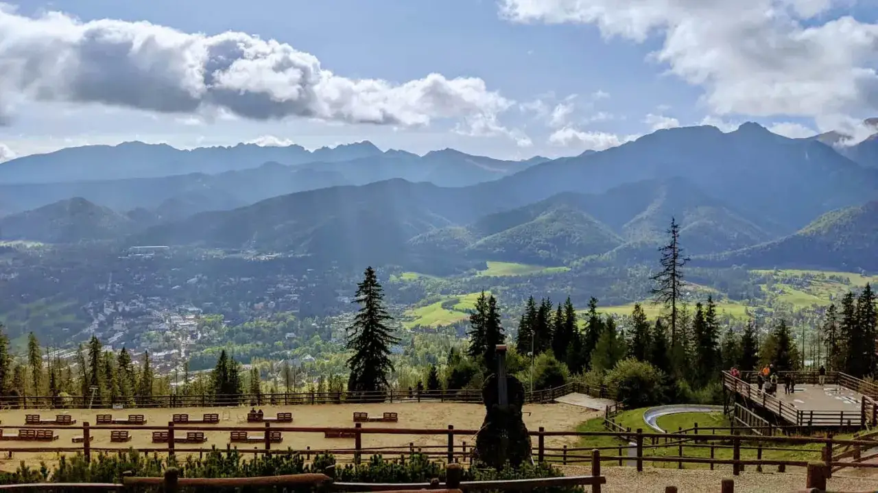 Widok na Tatry z Gubałówki. W oddali miasto, na pierwszym planie ławki i rzeźba. Zastanawiasz się, ile się idzie na Gubałówkę?