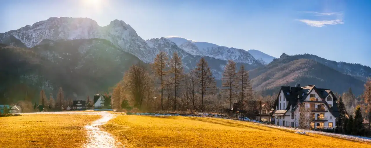 Widok z Giewontu na zaśnieżone szczyty i złote pola. W dole domki i drzewa.