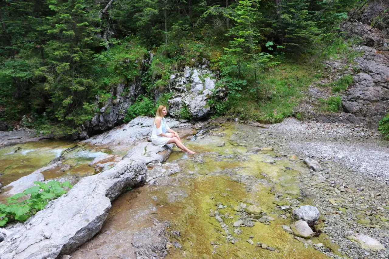 Kobieta w białej sukience siedzi na kamieniu nad strumieniem w Dolinie Białego, Zakopane. Wokół bujna zieleń i skaliste zbocza.